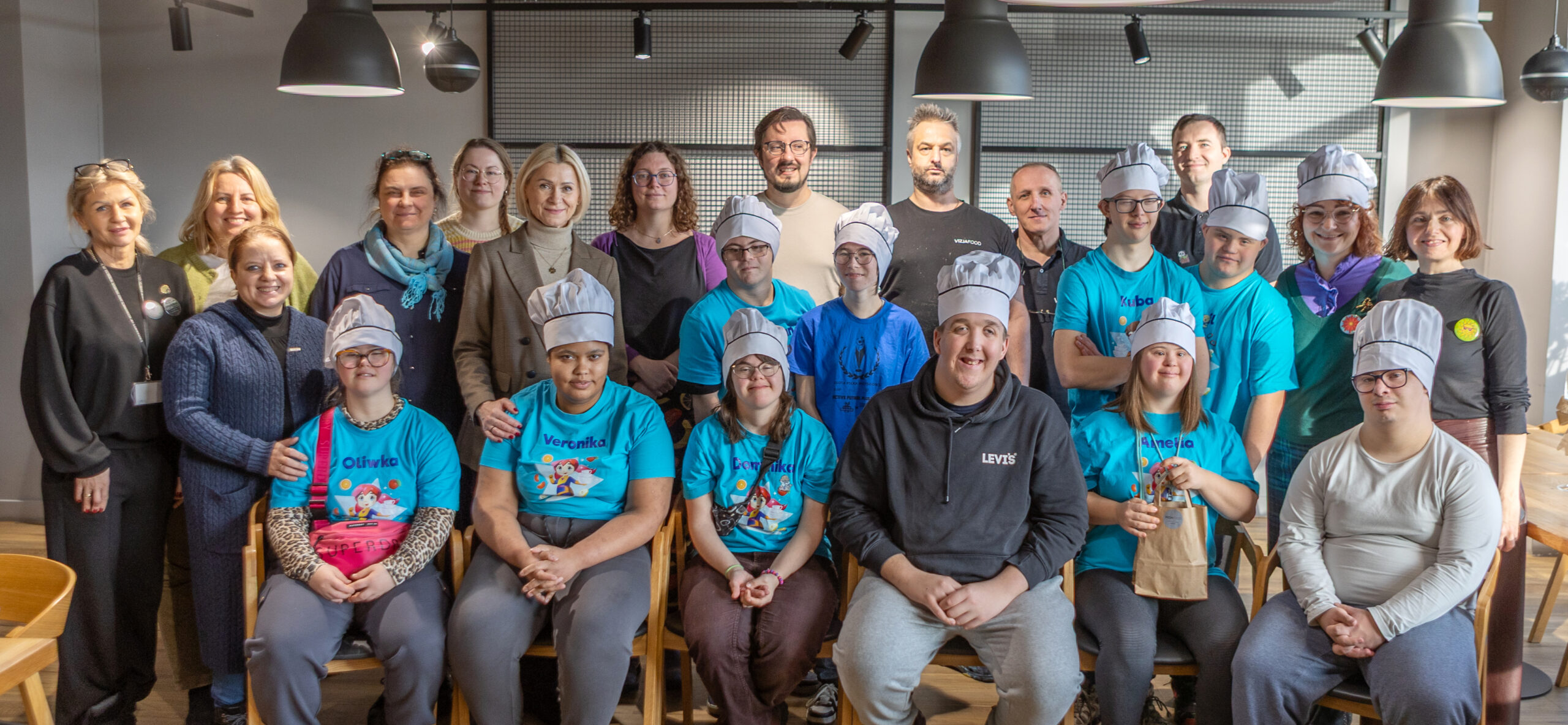 osoby z niepełnosprawnościami pozujące do zdjęcia grupowego po zajęciach przygotowujących do pracy w kuchni; osoby ubrane są w niebieskie bluzy i czapki kucharskie