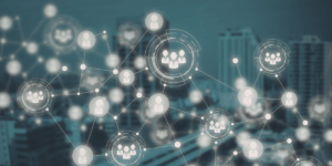 Grafika wyobrażająca sieć powiązań i relacji. Abstrakcyjna sieć linii łącząca jasne ikony przedstawiające sylwetki ludzi oraz grupy osób. Niektóre elementy są wyróżnione cyfrowymi okręgami przypominającymi interfejs analityczny. W tle widać rozmyte wieżowce w chłodnej, niebiesko-szarej tonacji.