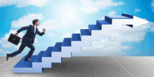 Mężczyzna w granatowym garniturze, trzymający teczkę, dynamicznie wbiega po schodach. Konstrukcja schodów ma kształt dużej, niebieskiej strzałki skierowanej ukośnie w prawy górny róg, na tle błękitnego nieba z chmurami. Ilustracja symbolizuje awans i dążenie do celu. Obrazuje rozwój kariery i sukces zawodowy.
