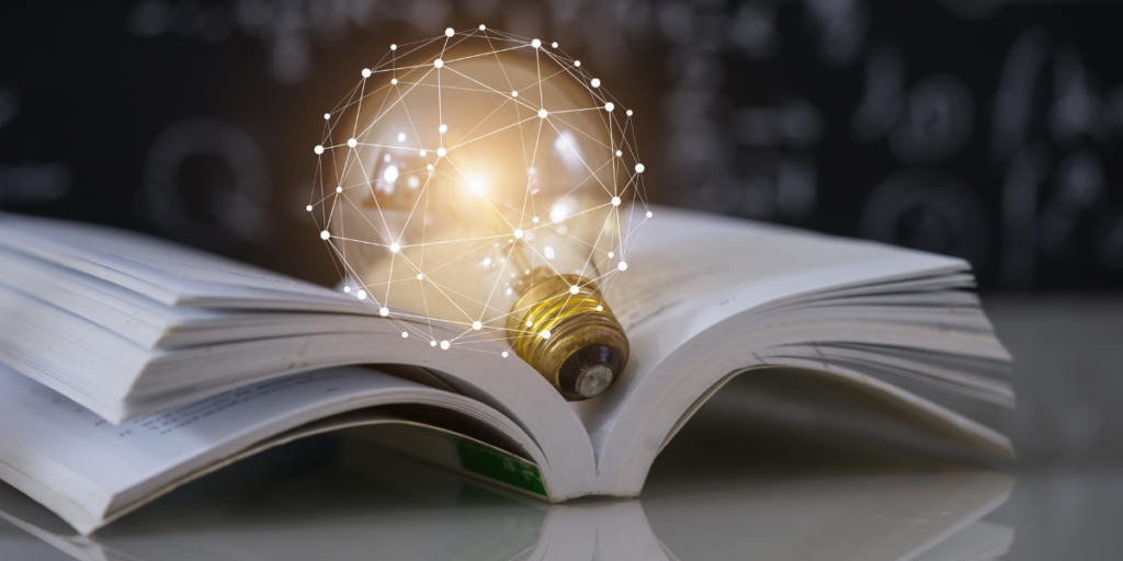 Symboliczna grafika przedstawiająca połączenie tradycyjnej nauki z nowoczesną technologią: otwarta książka, na której spoczywa świecąca żarówka opleciona cyfrową siecią białych linii i punktów. W tle widać ciemną tablicę szkolną z rozmytymi wzorami matematycznymi