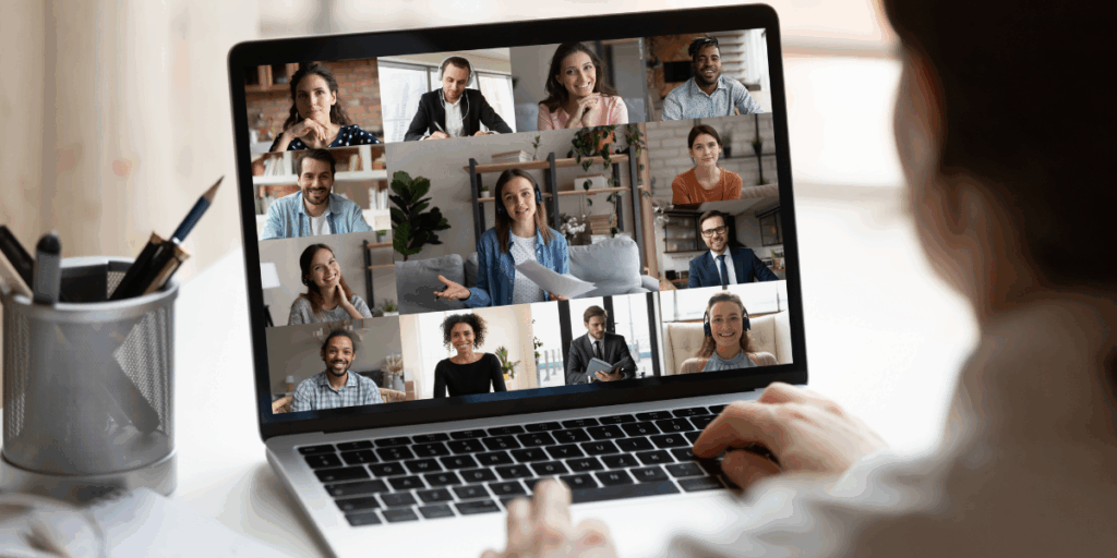 Widok zza ramienia osoby korzystającej z laptopa, na którego ekranie trwa wideokonferencja. Na monitorze wyświetla się galeria 12 uczestników (kobiet i mężczyzn) w różnych wnętrzach - domowych i biurowych. W centrum znajduje się większe okno z kobietą w słuchawkach, która gestykuluje i trzyma dokumenty, prowadząc prezentację. Pozostali uczestnicy patrzą w stronę kamer, uśmiechając się lub słuchając z uwagą. Na biurku obok laptopa stoi srebrny przybornik z długopisami. Zdjęcie jest ilustracją idei konsultacji online.