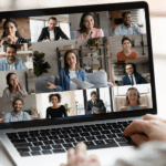 Widok zza ramienia osoby korzystającej z laptopa, na którego ekranie trwa wideokonferencja. Na monitorze wyświetla się galeria 12 uczestników (kobiet i mężczyzn) w różnych wnętrzach - domowych i biurowych. W centrum znajduje się większe okno z kobietą w słuchawkach, która gestykuluje i trzyma dokumenty, prowadząc prezentację. Pozostali uczestnicy patrzą w stronę kamer, uśmiechając się lub słuchając z uwagą. Na biurku obok laptopa stoi srebrny przybornik z długopisami. Zdjęcie jest ilustracją idei konsultacji online.