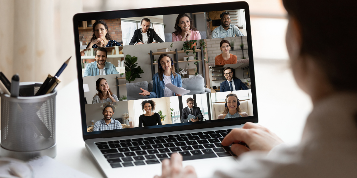 Widok zza ramienia osoby korzystającej z laptopa, na którego ekranie trwa wideokonferencja. Na monitorze wyświetla się galeria 12 uczestników (kobiet i mężczyzn) w różnych wnętrzach - domowych i biurowych. W centrum znajduje się większe okno z kobietą w słuchawkach, która gestykuluje i trzyma dokumenty, prowadząc prezentację. Pozostali uczestnicy patrzą w stronę kamer, uśmiechając się lub słuchając z uwagą. Na biurku obok laptopa stoi srebrny przybornik z długopisami. Zdjęcie jest ilustracją idei konsultacji online.