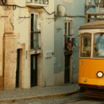 Zabytkowy żółty tramwaj linii 28, jadący wąską, brukowaną uliczką w Lizbonie obok starych kamienic z wiszącym na balkonach praniem.