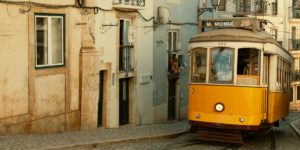 Zabytkowy żółty tramwaj linii 28, jadący wąską, brukowaną uliczką w Lizbonie obok starych kamienic z wiszącym na balkonach praniem.