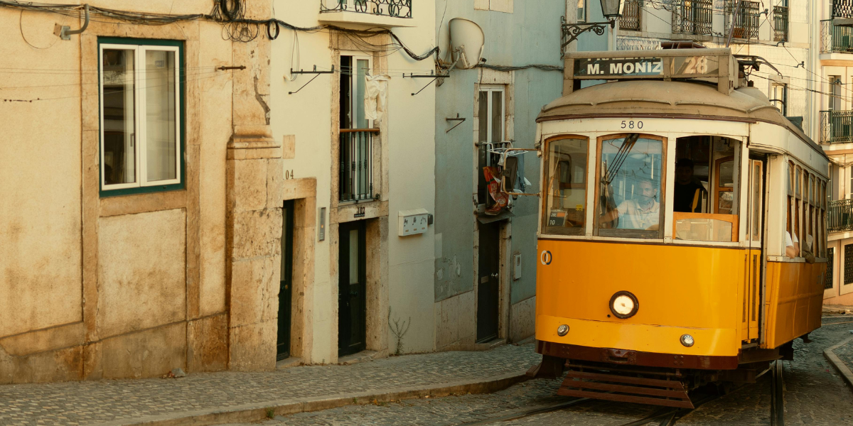 Zabytkowy żółty tramwaj linii 28, jadący wąską, brukowaną uliczką w Lizbonie obok starych kamienic z wiszącym na balkonach praniem.