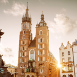 Widok na krakowski Rynek Główny o złotej godzinie. Na pierwszym planie pomnik Adama Mickiewicza, w tle monumentalna Bazylika Mariacka z dwiema wieżami oraz zabytkowe kamienice. Ciepłe światło słoneczne tworzy efektowny blask.