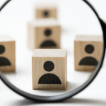 Zdjęcie przedstawiające rozrzucone na białym tle jasne, drewniane kostki z czarnymi ikonami sylwetek ludzi. Na pierwszym planie dłoń trzyma czarną lupę, która powiększa jedną z kostek, skupiając na niej uwagę.