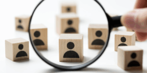 Zdjęcie przedstawiające rozrzucone na białym tle jasne, drewniane kostki z czarnymi ikonami sylwetek ludzi. Na pierwszym planie dłoń trzyma czarną lupę, która powiększa jedną z kostek, skupiając na niej uwagę.