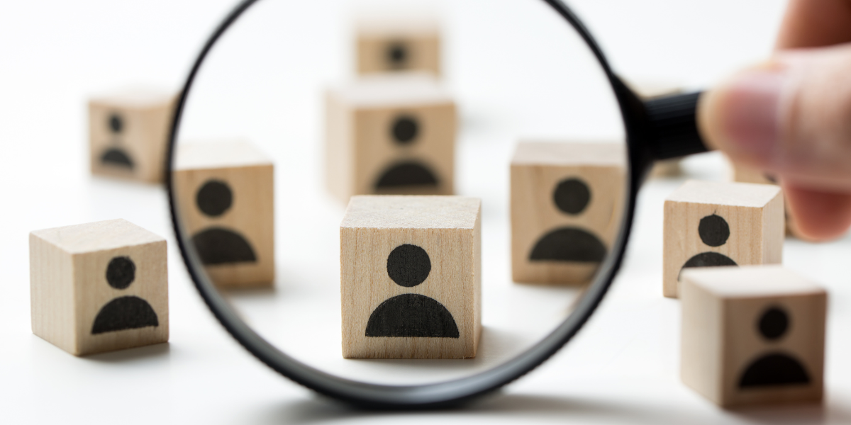 Zdjęcie przedstawiające rozrzucone na białym tle jasne, drewniane kostki z czarnymi ikonami sylwetek ludzi. Na pierwszym planie dłoń trzyma czarną lupę, która powiększa jedną z kostek, skupiając na niej uwagę.