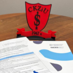 Program seminarium dotyczącego Zintegrowanego Systemu Kwalifikacji leżący na drewnianym stole obok czerwonej tarczy pamiątkowej z logo Centrum Kształcenia Zawodowego i Ustawicznego w Strzelinie i datą 1947.