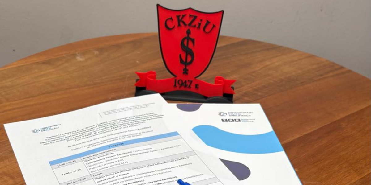 Program seminarium dotyczącego Zintegrowanego Systemu Kwalifikacji leżący na drewnianym stole obok czerwonej tarczy pamiątkowej z logo Centrum Kształcenia Zawodowego i Ustawicznego w Strzelinie i datą 1947.