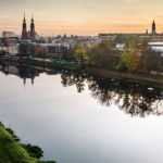 Malownicza panorama Opola widziana znad Odry podczas złotego zachodu słońca. Na horyzoncie dominują wieże katedry oraz charakterystyczna Wieża Piastowska. W tafli spokojnej rzeki odbijają się budynki miasta, drzewa w jesiennych barwach oraz nowoczesny most pieszo-rowerowy.
