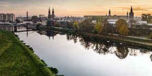 Malownicza panorama Opola widziana znad Odry podczas złotego zachodu słońca. Na horyzoncie dominują wieże katedry oraz charakterystyczna Wieża Piastowska. W tafli spokojnej rzeki odbijają się budynki miasta, drzewa w jesiennych barwach oraz nowoczesny most pieszo-rowerowy.