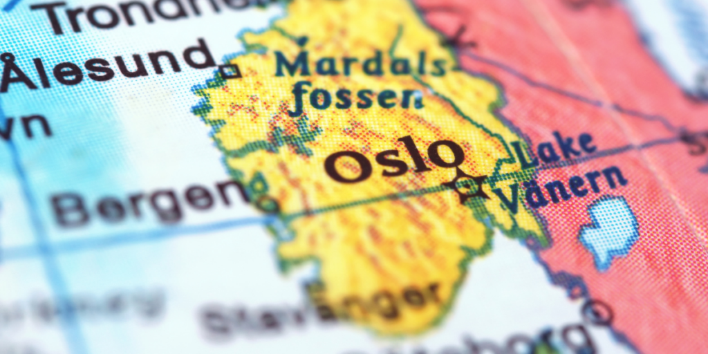 Zdjęcie typu makro przedstawiające fragment kolorowej, drukowanej mapy Skandynawii. Centralnym punktem kadru jest żółty obszar z wyraźnym, wyostrzonym napisem „Oslo” oznaczonym symbolem gwiazdy. Otoczenie ma niską głębię ostrości – napisy wokół, takie jak „Bergen”, „Stavanger”, „Mardalsfossen” oraz „Lake Vänern”, są lekko rozmyte. W lewym górnym rogu widoczny jest fragment napisu „Ålesund”.