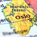 Zdjęcie typu makro przedstawiające fragment kolorowej, drukowanej mapy Skandynawii. Centralnym punktem kadru jest żółty obszar z wyraźnym, wyostrzonym napisem „Oslo” oznaczonym symbolem gwiazdy. Otoczenie ma niską głębię ostrości – napisy wokół, takie jak „Bergen”, „Stavanger”, „Mardalsfossen” oraz „Lake Vänern”, są lekko rozmyte. W lewym górnym rogu widoczny jest fragment napisu „Ålesund”.