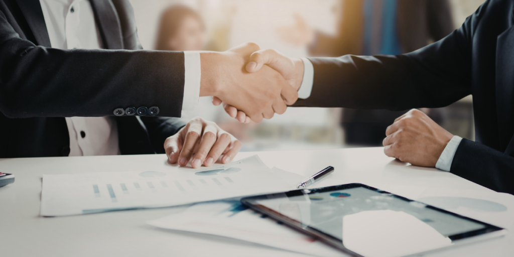 Zdjęcie wyobrażające zawarcie umowy: zbliżenie na uścisk dłoni dwóch osób w strojach biznesowych, siedzących przy białym stole z wykresami, dokumentami i tabletem. W tle widoczne rozmyte biuro