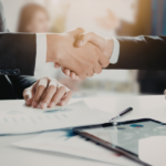 Zdjęcie wyobrażające zawarcie umowy: zbliżenie na uścisk dłoni dwóch osób w strojach biznesowych, siedzących przy białym stole z wykresami, dokumentami i tabletem. W tle widoczne rozmyte biuro