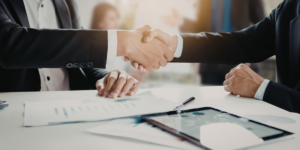 Zdjęcie wyobrażające zawarcie umowy: zbliżenie na uścisk dłoni dwóch osób w strojach biznesowych, siedzących przy białym stole z wykresami, dokumentami i tabletem. W tle widoczne rozmyte biuro