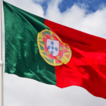 Flaga Portugalii, zielono-czerwona z tradycyjnym godłem pośrodku, powiewająca na białym maszcie na tle błękitnego nieba z białymi chmurami.
