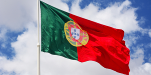 Flaga Portugalii, zielono-czerwona z tradycyjnym godłem pośrodku, powiewająca na białym maszcie na tle błękitnego nieba z białymi chmurami.