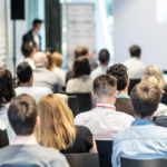 Widok z perspektywy widowni na grupę osób biorących udział w seminarium. Uczestnicy siedzą w rzędach, zwróceni w stronę rozmytej sylwetki prelegenta i ekranu projekcyjnego na przodzie jasnej sali.