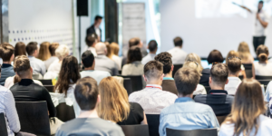 Widok z perspektywy widowni na grupę osób biorących udział w seminarium. Uczestnicy siedzą w rzędach, zwróceni w stronę rozmytej sylwetki prelegenta i ekranu projekcyjnego na przodzie jasnej sali.