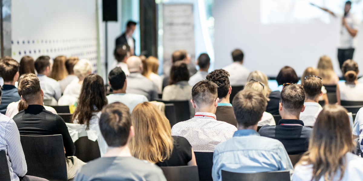 Widok z perspektywy widowni na grupę osób biorących udział w seminarium. Uczestnicy siedzą w rzędach, zwróceni w stronę rozmytej sylwetki prelegenta i ekranu projekcyjnego na przodzie jasnej sali.
