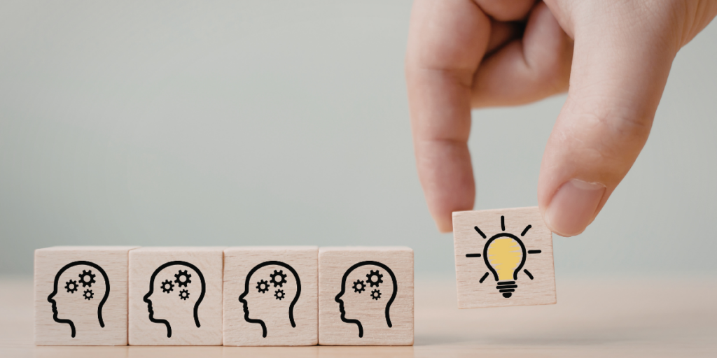 Dłoń dokładająca drewniany klocek z ikoną zapalonej żarówki do rzędu klocków z symbolami ludzkich głów i kół zębatych - symboliczna ilustracja procesu przekazywania idei i zaszczepiania pomysłów.