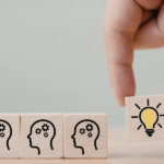 Dłoń dokładająca drewniany klocek z ikoną zapalonej żarówki do rzędu klocków z symbolami ludzkich głów i kół zębatych - symboliczna ilustracja procesu przekazywania idei i zaszczepiania pomysłów.