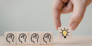 Dłoń dokładająca drewniany klocek z ikoną zapalonej żarówki do rzędu klocków z symbolami ludzkich głów i kół zębatych - symboliczna ilustracja procesu przekazywania idei i zaszczepiania pomysłów.