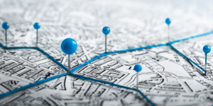 Artystyczne zdjęcie starej mapy z zaznaczonymi punktami. Sześć niebieskich pinezek o kulistych główkach jest wbitych w różne miejsca mapy, a między nimi rozciągnięty jest jaskrawoniebieski sznurek. Główna pinezka znajduje się na pierwszym planie, lekko po lewej stronie, stanowiąc centralny węzeł trasy. Tło mapy jest rozmyte, co nadaje zdjęciu głębi. Metaforyczne przedstawienie planowania trasy lub sieci połączeń.