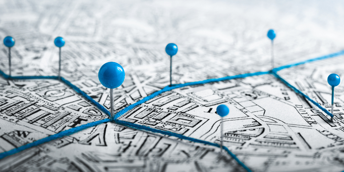 Artystyczne zdjęcie starej mapy z zaznaczonymi punktami. Sześć niebieskich pinezek o kulistych główkach jest wbitych w różne miejsca mapy, a między nimi rozciągnięty jest jaskrawoniebieski sznurek. Główna pinezka znajduje się na pierwszym planie, lekko po lewej stronie, stanowiąc centralny węzeł trasy. Tło mapy jest rozmyte, co nadaje zdjęciu głębi. Metaforyczne przedstawienie planowania trasy lub sieci połączeń.