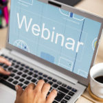 dłonie osoby piszącej na laptopie, na wyświetlaczu którego jest napis "webinar"