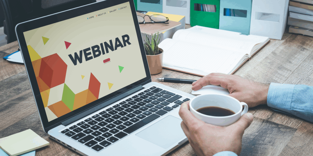 Zdjęcie przedstawiające osobę przy biurku z otwartym laptopem, na którego ekranie widnieje duży napis »WEBINAR« na tle kolorowych figur geometrycznych. Jedna dłoń spoczywa na myszce, a druga trzyma biały kubek z czarną kawą. Na drewnianym blacie znajdują się również okulary, notatnik, długopis i mały kaktus.