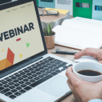 Zdjęcie przedstawiające osobę przy biurku z otwartym laptopem, na którego ekranie widnieje duży napis »WEBINAR« na tle kolorowych figur geometrycznych. Jedna dłoń spoczywa na myszce, a druga trzyma biały kubek z czarną kawą. Na drewnianym blacie znajdują się również okulary, notatnik, długopis i mały kaktus.