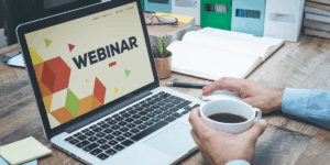 Zdjęcie przedstawiające osobę przy biurku z otwartym laptopem, na którego ekranie widnieje duży napis »WEBINAR« na tle kolorowych figur geometrycznych. Jedna dłoń spoczywa na myszce, a druga trzyma biały kubek z czarną kawą. Na drewnianym blacie znajdują się również okulary, notatnik, długopis i mały kaktus.