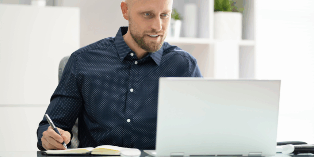 Zdjęcie ilustrujące uczestnika webinaru podczas aktywnego robienia notatek. Mężczyzna z krótkim zarostem i w ciemnej koszuli w drobne białe kropki siedzi przy biurku, pracując przy laptopie. Z uśmiechem patrzy w ekran komputera, a jednocześnie prawą ręką robi notatki w otwartym zeszycie. Tło jest jasne i nowoczesne, z widocznymi regałami i roślinami doniczkowymi, co sugeruje przestrzeń biurową lub domowy gabinet.