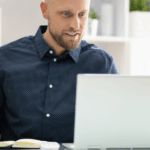 Zdjęcie ilustrujące uczestnika webinaru podczas aktywnego robienia notatek. Mężczyzna z krótkim zarostem i w ciemnej koszuli w drobne białe kropki siedzi przy biurku, pracując przy laptopie. Z uśmiechem patrzy w ekran komputera, a jednocześnie prawą ręką robi notatki w otwartym zeszycie. Tło jest jasne i nowoczesne, z widocznymi regałami i roślinami doniczkowymi, co sugeruje przestrzeń biurową lub domowy gabinet.