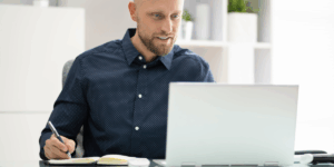Zdjęcie ilustrujące uczestnika webinaru podczas aktywnego robienia notatek. Mężczyzna z krótkim zarostem i w ciemnej koszuli w drobne białe kropki siedzi przy biurku, pracując przy laptopie. Z uśmiechem patrzy w ekran komputera, a jednocześnie prawą ręką robi notatki w otwartym zeszycie. Tło jest jasne i nowoczesne, z widocznymi regałami i roślinami doniczkowymi, co sugeruje przestrzeń biurową lub domowy gabinet.