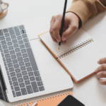 osoba pisząca w notesie obok laptopa